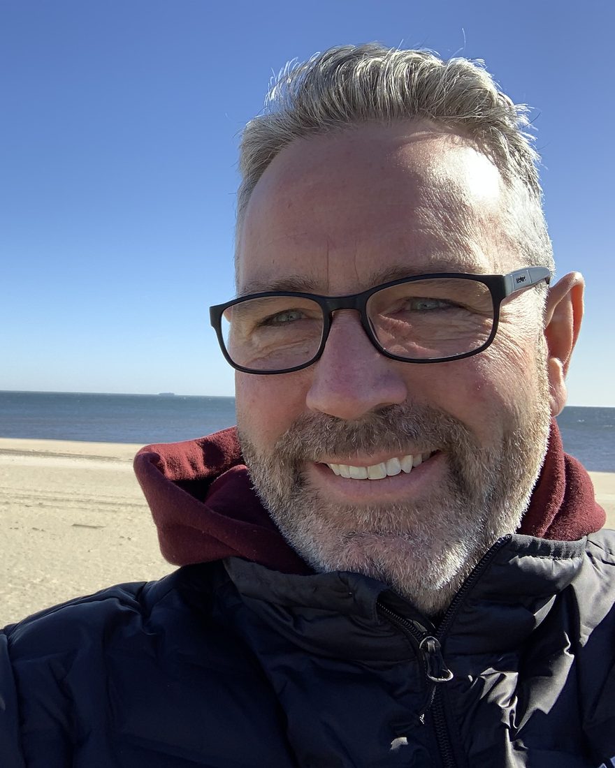 Chad Gillikin smiling on a sunny beach, wearing glasses, a dark jacket, and a maroon hoodie, with the ocean behind him.